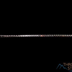 May include: A silver-colored box chain necklace against a black background. The chain is composed of small, square links, creating a sleek and modern design. The necklace is a jewelry item.