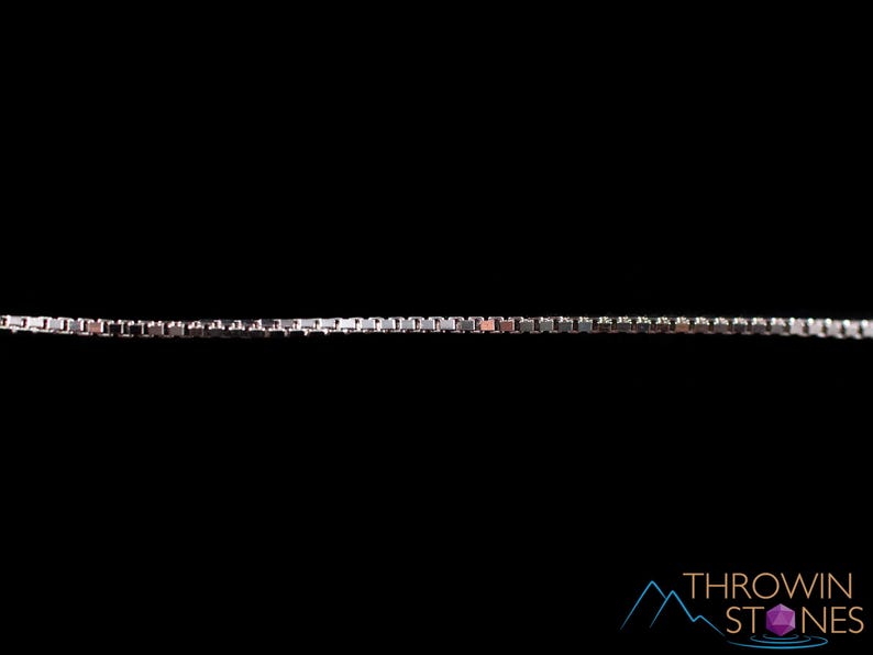 May include: A silver-colored box chain necklace against a black background. The chain is composed of small, square links, creating a sleek and modern design. The necklace is a jewelry item.
