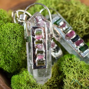 This crystal pendant features a clear Quartz gemstone, showcasing its transparent beauty and amplifying properties. Surrounding the Quartz are seven, green and pink Tourmaline gemstone accent crystals.