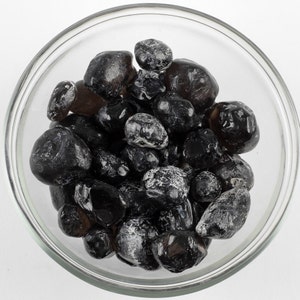 May include: A clear glass bowl filled with dark, smooth, rounded stones. The stones vary in size and have a glossy, reflective surface. Some stones have a frosted, white appearance. The bowl is set against a plain white background.