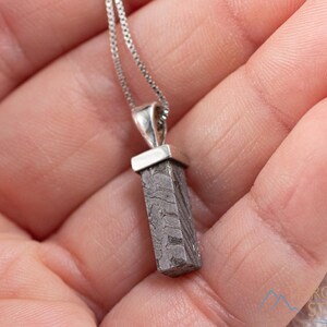 May include: A silver necklace with a rectangular, patterned pendant. The pendant is a dark gray color with a unique, textured design. The necklace is held in a person's hand.