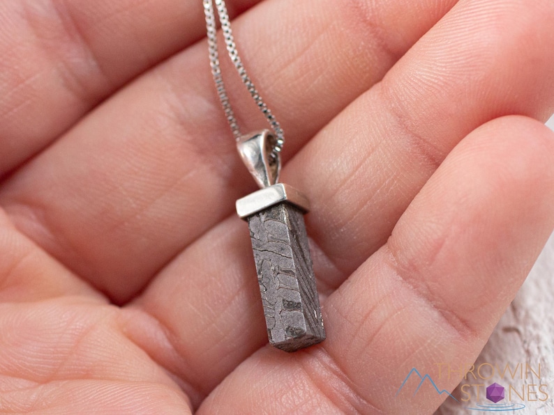 May include: A silver necklace with a rectangular, patterned pendant. The pendant is a dark gray color with a unique, textured design. The necklace is held in a person's hand.
