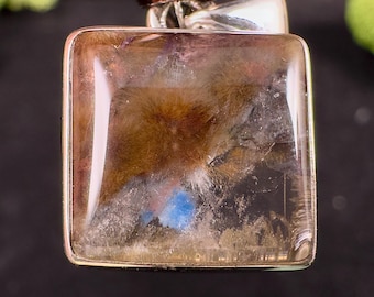 May include: A square pendant with a silver-colored metal frame. The pendant features a translucent, multi-colored stone with brown, blue, and white inclusions. The stone has a polished, glossy surface. The pendant is likely for a necklace.