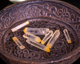 May include: A collection of small, clear quartz crystals with a yellow tint, resting in a dark brown bowl with an intricate design.