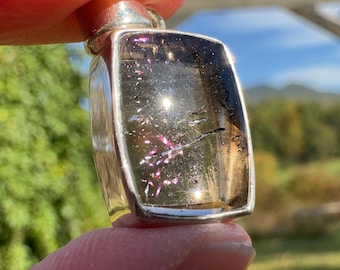 May include: A silver pendant with a clear, faceted gemstone. The gemstone has a light brown color with pink and silver inclusions. The pendant is rectangular in shape and has a smooth, polished finish.