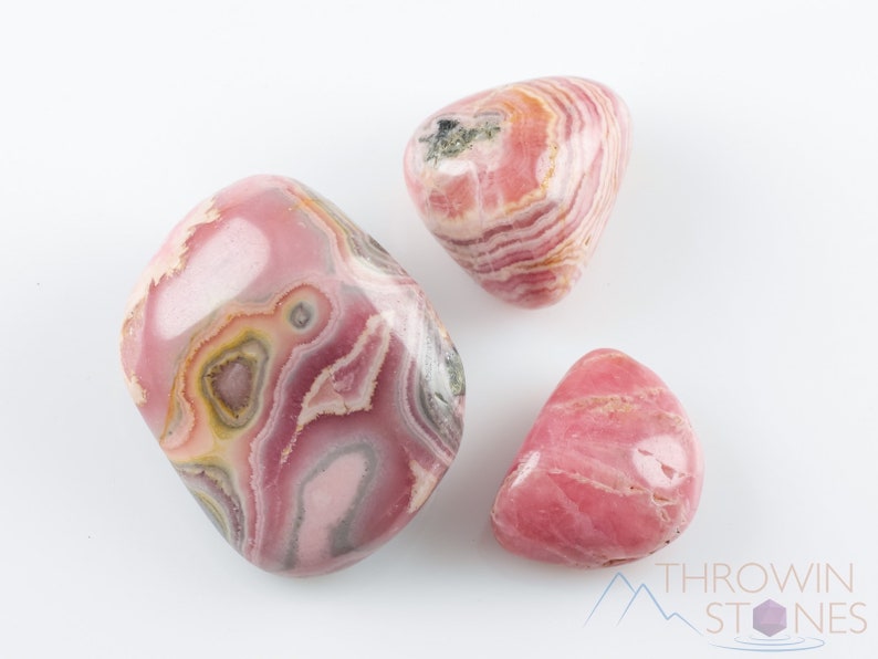 Rhodochrosite Tumbled Stones. These polished crystals are pink with yellowish grey white and brown bands. Each crystal is unique in color and you will receive 1 stone that is 0.7 to 1.7 inches long and weighs 6 to 65 grams. Listing has variations.