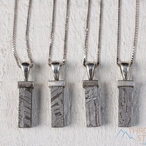 May include: Four silver necklaces with rectangular, patterned pendants. The pendants are a gray, textured material, possibly stone or metal. Each necklace has a delicate silver chain and a simple, elegant design. The background is a white, textured surface.