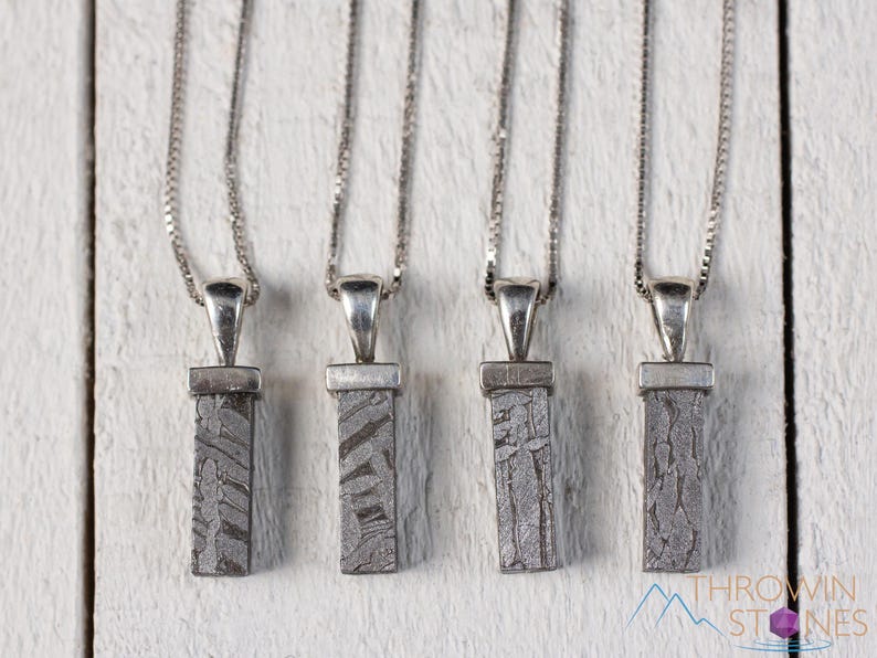 May include: Four silver necklaces with rectangular, patterned pendants. The pendants are a gray, textured material, possibly stone or metal. Each necklace has a delicate silver chain and a simple, elegant design. The background is a white, textured surface.