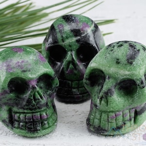 May include: Three green, black and purple gemstone skull carvings. The skulls are carved from a polished stone with a mottled appearance.