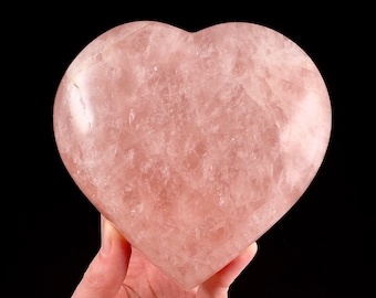 Puede incluir: Un gran cristal de cuarzo rosa en forma de corazón. El cristal es de color rosa pálido y tiene una superficie lisa y pulida.