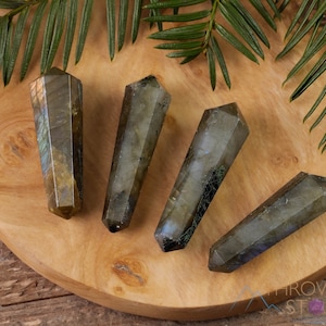 May include: Four iridescent labradorite crystal wands, each with a pointed tip, are arranged on a wooden surface. The crystals are a dark gray with flashes of blue, green, and gold.