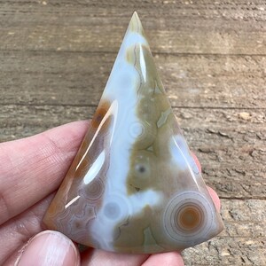 May include: A triangular shaped agate cabochon with a white, brown, and tan color pattern. The agate has a smooth, polished surface.