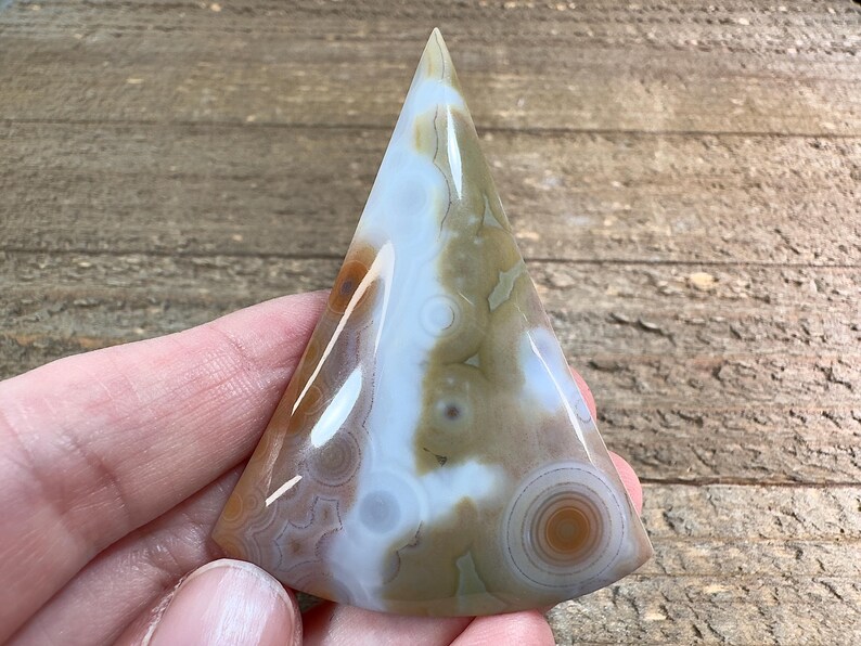 May include: A triangular shaped agate cabochon with a white, brown, and tan color pattern. The agate has a smooth, polished surface.