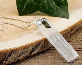 May include: A rectangular, clear selenite crystal pendant with a silver-colored metal cap and a small green stone accent, suspended from a silver chain. The pendant is displayed on a wooden surface with a green leaf in the background.