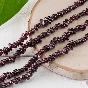 May include: A strand of dark red garnet chip beads, displayed on a wooden surface. The beads are small and irregular in shape, with a natural, unpolished finish.