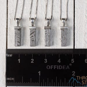 May include: Four silver pendant necklaces with rectangular, patterned meteorite pendants. The pendants are suspended from delicate silver chains. A ruler with inch and millimeter markings is in the foreground. The background is a white, textured surface.