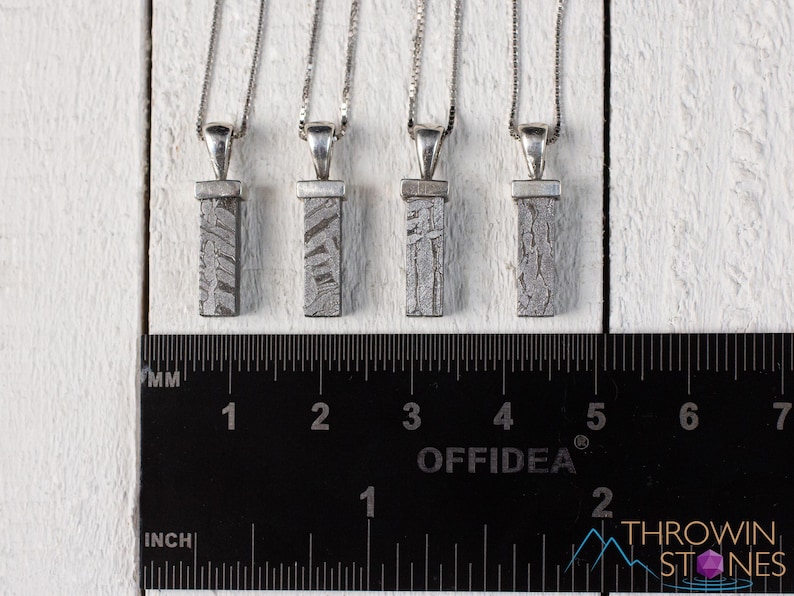 May include: Four silver pendant necklaces with rectangular, patterned meteorite pendants. The pendants are suspended from delicate silver chains. A ruler with inch and millimeter markings is in the foreground. The background is a white, textured surface.
