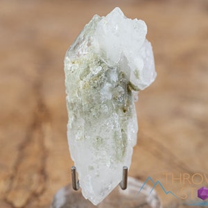 Puede incluir: Un cristal de cuarzo transparente con inclusiones verdes. El cristal está en un soporte y tiene una punta puntiaguda.