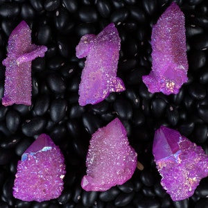 May include: Six iridescent purple amethyst crystals with a sparkly, textured surface, arranged on a bed of black beans.