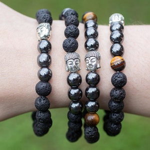 Puede incluir: Cuatro pulseras de cuentas apiladas en una muñeca. Las pulseras están hechas de cuentas de lava negra, cuentas de hematita negra y cuentas de ojo de tigre. Cada pulsera tiene un dije de Buda plateado.