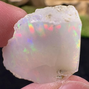 May include: A rough, white opal gemstone with iridescent flashes of color. The opal is held between two fingers, showcasing its natural, unpolished texture and the play of light across its surface. The colors include pink, green, and yellow.