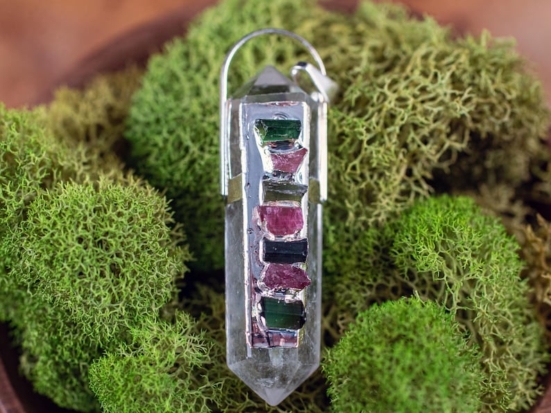 This crystal pendant features a clear Quartz gemstone, showcasing its transparent beauty and amplifying properties. Surrounding the Quartz are seven, green and pink Tourmaline gemstone accent crystals.