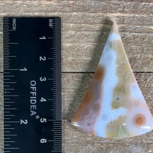 May include: A triangular shaped agate cabochon with a white, brown, and orange color pattern. The cabochon is approximately 2 inches in length.