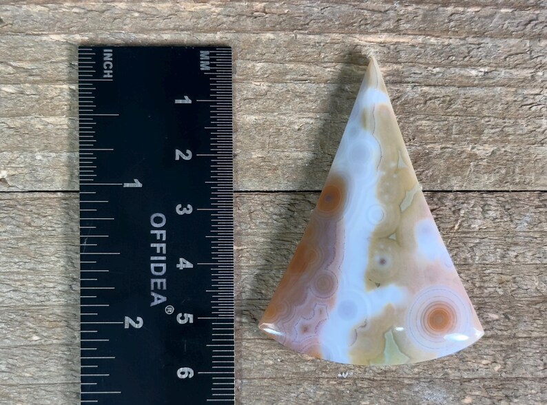 May include: A triangular shaped agate cabochon with a white, brown, and orange color pattern. The cabochon is approximately 2 inches in length.