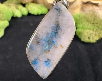 May include: A gemstone pendant with a silver bail. The pendant features a light blue and white stone with blue and brown inclusions. The stone is polished and has a smooth surface. The pendant is a unique piece of jewelry.