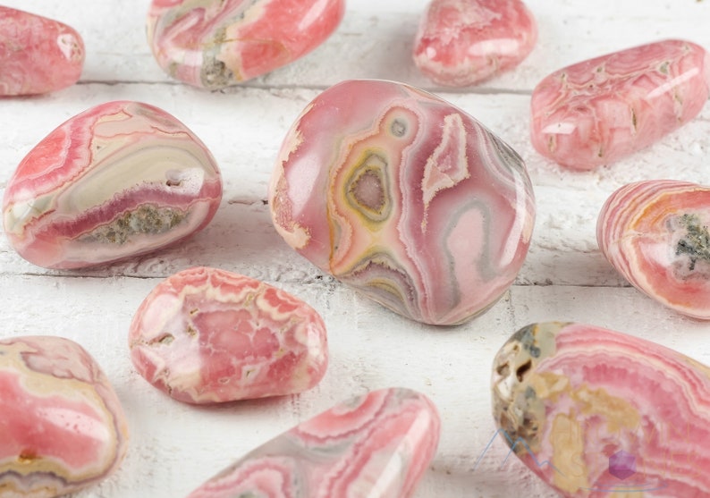 Rhodochrosite Tumbled Stones. These polished crystals are pink with yellowish grey white and brown bands. Each crystal is unique in color and you will receive 1 stone that is 0.7 to 1.7 inches long and weighs 6 to 65 grams. Listing has variations.