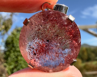 Op de afbeelding: Een ronde, rode en heldere kristallen hanger met zilveren accenten. De kristal heeft een uniek, draaiend patroon van rode en heldere insluitsels.