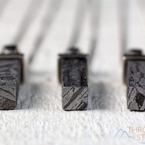 May include: Close-up of three rectangular, dark gray objects with a textured, patterned surface. The objects are set against a blurred white background. The objects appear to be made of stone or metal. The bottom right corner of the image has the text "THROWIN STONES".