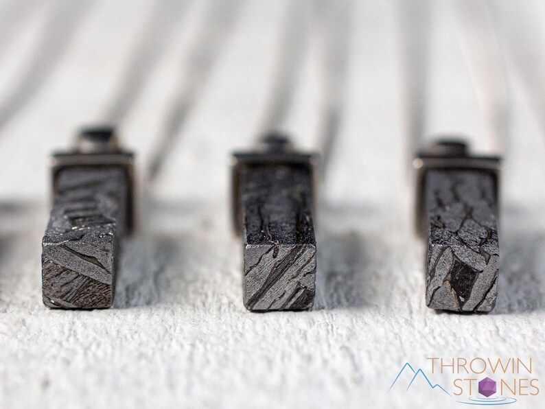 May include: Close-up of three rectangular, dark gray objects with a textured, patterned surface. The objects are set against a blurred white background. The objects appear to be made of stone or metal. The bottom right corner of the image has the text "THROWIN STONES".