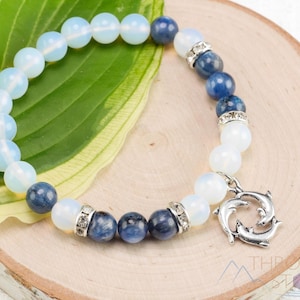 May include: A bracelet with light blue and dark blue beads, with silver spacers. A silver charm with two dolphins hangs from the bracelet. The bracelet is displayed on a wooden surface with a green leaf.