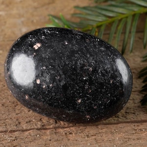 May include: A smooth, black obsidian palm stone with a speckled, silver-grey pattern. The stone is resting on a wooden surface.