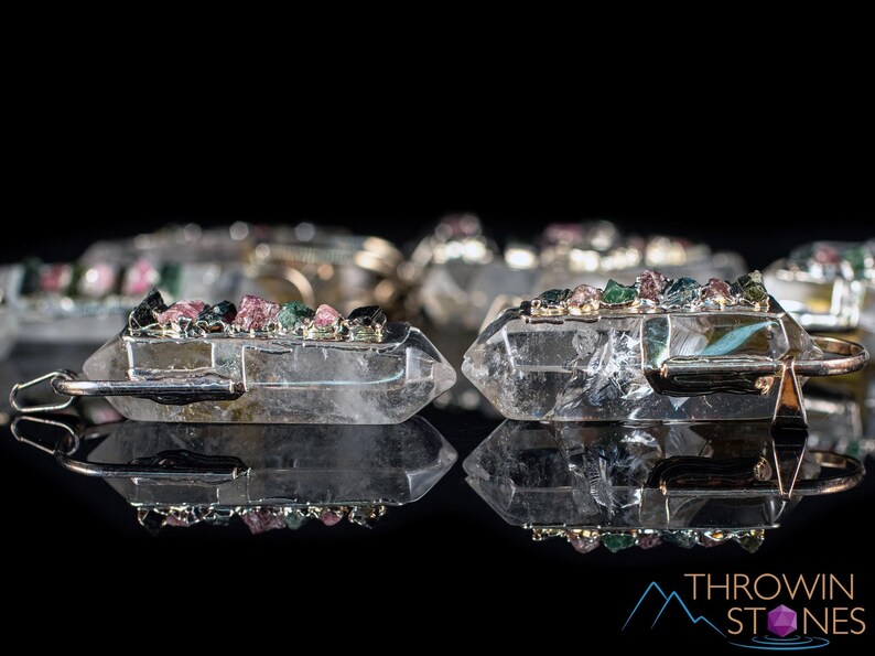 This crystal pendant features a clear Quartz gemstone, showcasing its transparent beauty and amplifying properties. Surrounding the Quartz are seven, green and pink Tourmaline gemstone accent crystals.