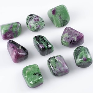 Puede incluir: Nueve piedras pulidas de zoisita verde y púrpura, una variedad de granate grossular verde, sobre un fondo blanco.