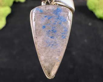 May include: A silver pendant with a triangular, polished stone. The stone has a translucent, light gray base with blue, brushstroke-like inclusions. The pendant is suspended from a silver bail, showcasing a unique, natural gemstone.