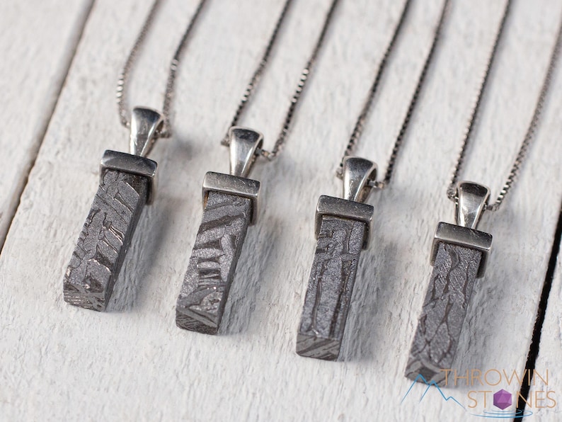 May include: Four rectangular pendants with a textured, dark gray pattern are displayed on silver chains. Each pendant is attached to a silver bail. The pendants are arranged on a white wooden surface. The logo "Thrown Stones" is visible in the bottom right corner.