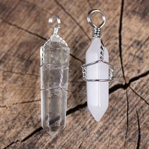 Könnte beinhalten: Zwei Kristallspitzen-Anhänger, die mit Silberdraht umwickelt sind. Der Anhänger links ist aus klarem Quarz, der Anhänger rechts ist weiß.