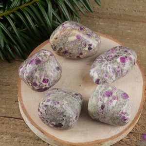 May include: Five tumbled stones with a white base and pink and purple flecks. The stones are arranged on a wooden surface.