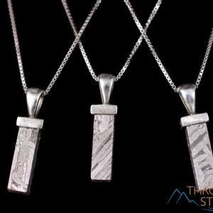 May include: Three silver necklaces with rectangular pendants. Each pendant has a textured, gray surface and hangs from a delicate chain. The background is black, highlighting the jewelry's design. The pendants appear to be made of a stone.