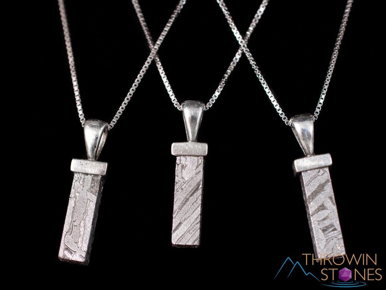 May include: Three silver necklaces with rectangular pendants. Each pendant has a textured, gray surface and hangs from a delicate chain. The background is black, highlighting the jewelry's design. The pendants appear to be made of a stone.