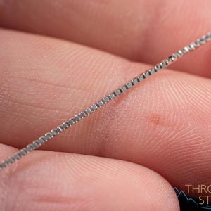 May include: A close-up of a silver box chain necklace. The chain is held in a person's hand, with the chain angled across the frame. The chain is made of small, square links. The background is a blurred image of skin.