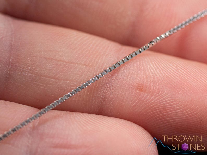 May include: A close-up of a silver box chain necklace. The chain is held in a person's hand, with the chain angled across the frame. The chain is made of small, square links. The background is a blurred image of skin.