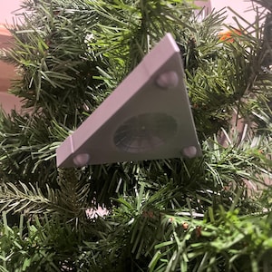 Puede incluir: Un accesorio de iluminación triangular gris para árbol de Navidad con tres pequeños pies blancos. El accesorio se coloca sobre un fondo de ramas verdes artificiales de árbol de Navidad.