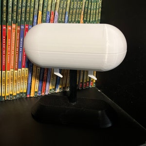 May include: White 3D printed airpod case with a black stand. The case is shaped like a blimp and has a ribbed texture. The stand has a rounded base.