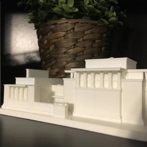 May include: A white 3D printed model of a building with a flat roof and a series of columns along the front.