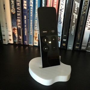 May include: A black Apple TV remote control is sitting on a white, 3D printed stand shaped like an apple. The stand is on a black surface.