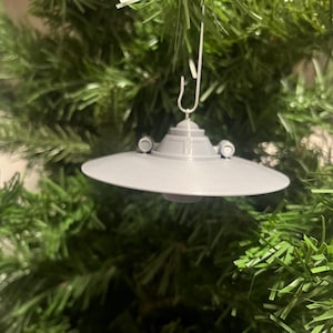 May include: A gray, 3D-printed UFO Christmas ornament. The ornament is suspended from a silver hook and features a classic flying saucer design with a circular base and two small, round protrusions. It hangs against a backdrop of green Christmas tree branches.